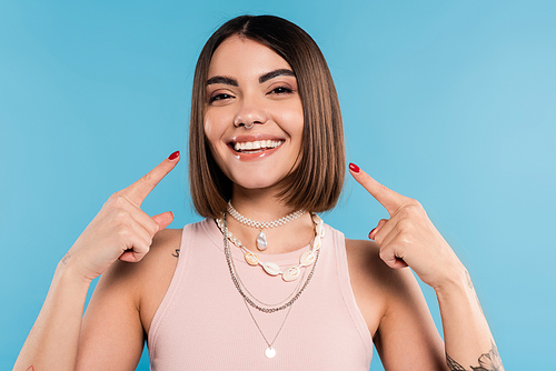 excitement, tattooed young woman with short brunette hair in tank top smiling and pointing at her cheeks on blue background, casual attire, gen z fashion, happiness, joyful