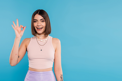 excitement, tattooed young woman with short hair in tank top smiling and showing ok gesture on blue background, casual attire, gen z fashion, fashionable trend, happiness