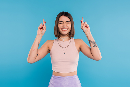 fingers crossed, tattooed young woman with short brunette hair in tank top making wish on blue background, casual attire, gen z fashion, happiness, smiling with closed eyes