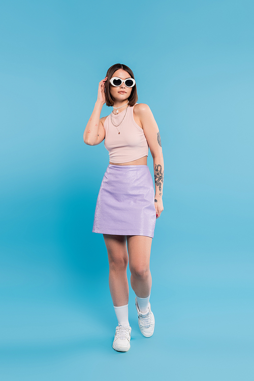 fashion trend, brunette young woman with short hair in tank top, skirt, white sneakers and sunglasses posing on blue background, casual attire, gen z fashion, personal style, nose piercing