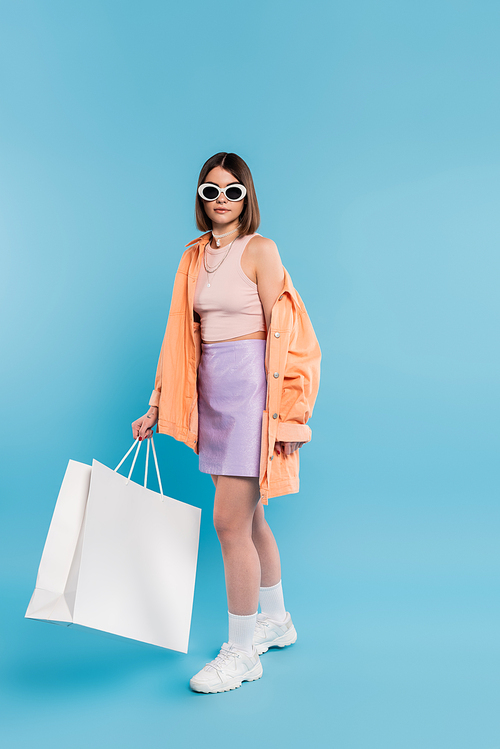 summer shopping, brunette young woman in tank top, skirt, sunglasses and orange shirt posing with shopping bag on blue background, casual attire, stylish posing, gen z, modern fashion