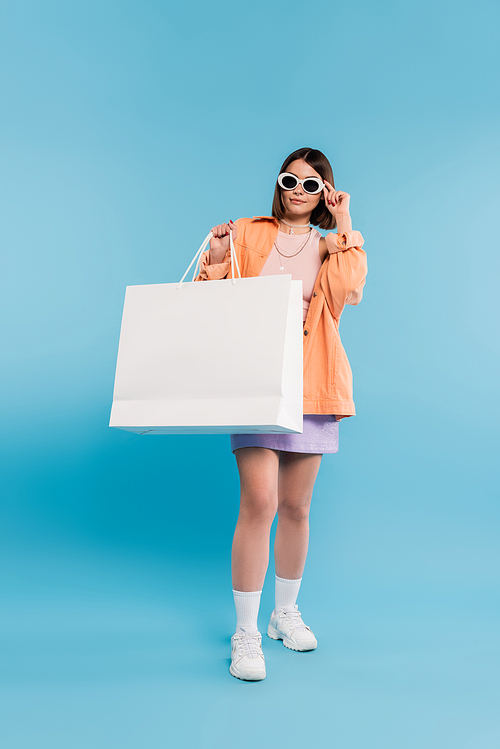 shopping spree, brunette young woman in tank top, skirt, sunglasses and orange shirt posing with shopping bag on blue background, casual attire, stylish posing, gen z, modern fashion