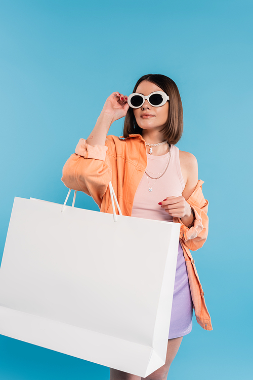 summer shopping, fashionable young woman in tank top, skirt, sunglasses and orange shirt posing with shopping bag on blue background, casual attire, stylish posing, gen z, modern fashion