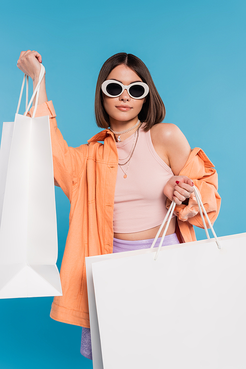 summer shopping, generation z, brunette young woman in tank top, skirt, sunglasses and orange shirt posing with shopping bags on blue background, casual attire, stylish posing, modern fashion