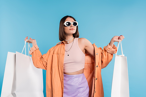 summer spree, brunette young woman in sunglasses and trendy outfit posing with shopping bags on blue background, casual attire, stylish posing, generation z, modern fashion