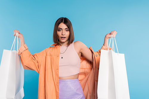 summer trends, pierced young woman in fashionable outfit posing with shopping bags on blue background, casual attire, stylish, generation z, modern fashion, orange shirt, tank top and skirt