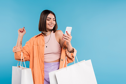 positivity, shopping spree, cheerful young woman in trendy outfit holding shopping bags and using smartphone on blue background, casual attire, stylish, generation z, modern fashion