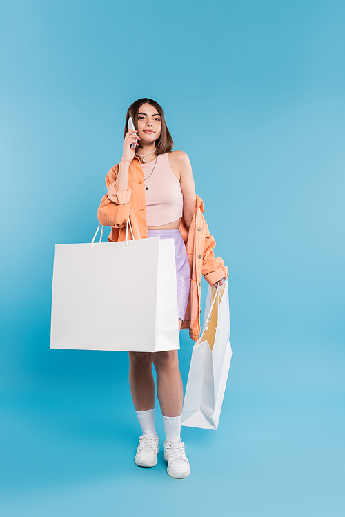 positivity, shopping spree, phone call, cheerful young woman in trendy outfit holding shopping bags and talking on smartphone on blue background, casual attire, generation z, modern fashion