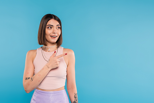 amazed woman showing something and looking away, stylish and tattooed model with nose piercing pointing with finger on blue background, generation z, summer fashion, smile