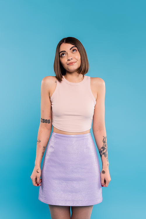 not knowing, smiling young woman with tattoos and nose piercing standing in tank top and skirt on blue background, looking up, confused, pretty face, generation z, summer outfit