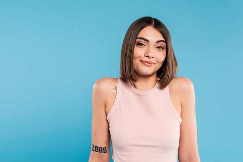not knowing, smiling young woman with tattoos and nose piercing standing in tank top on blue background, looking at camera, confused, pretty face, generation z, summer outfit