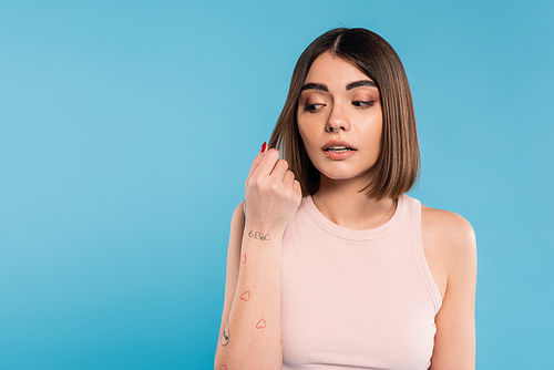hair damage, stress, concerned brunette young woman touching her hair on blue background, tattooed, nose piercing, tank top, generation z, casual attire, thinning hair