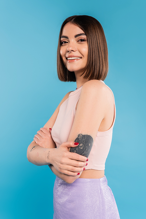folded hands, cheerful young woman with short hair and tattoos standing and looking at camera on blue background, nose piercing, youth, generation z, summer fashion
