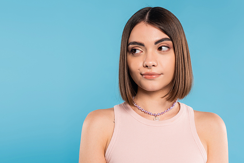 doubting, young brunette woman with short hair and nose piercing standing in tank top and looking away on blue background, youth culture, skepticism, generation z, summer fashion