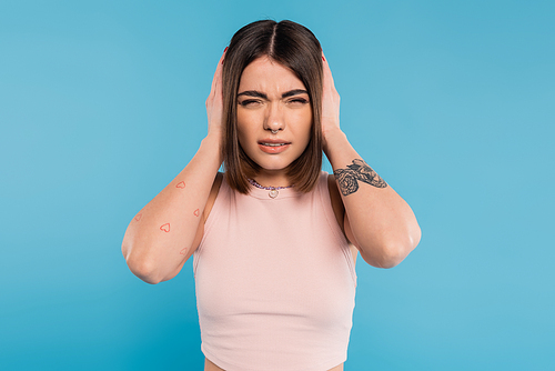 migraine, tattooed young woman with nose piercing and short hair touching head while suffering from headache on blue background, generation z, tiredness, stress