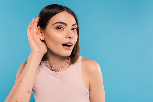 Don't hear you gesture, tattooed young woman with nose piercing and short hair holding hand near ear on blue background, generation z, listening, opened mouth, curious