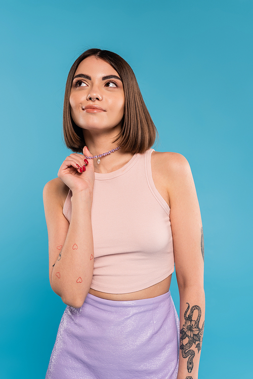 dreamy, tattooed young woman with nose piercing and short hair touching silver necklace and looking away on blue background, generation z, fashionable, trendy summer fashion