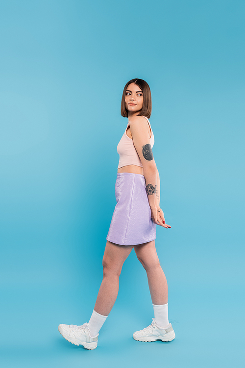 fashion trend, brunette young woman with short hair in tank top, skirt and white sneakers walking on blue background, casual attire, gen z fashion, personal style, nose piercing, everyday makeup