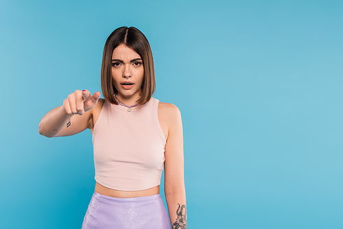 pointing at camera, emotional young woman with short hair, tattoos and pose piercing gesturing on blue background, generation z, displeased, casual attire, everyday makeup