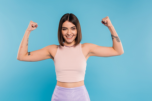 women power, cheerful young woman with short hair, tattoos and nose piercing showing muscles on blue background, generation z, displeased, casual attire, strength