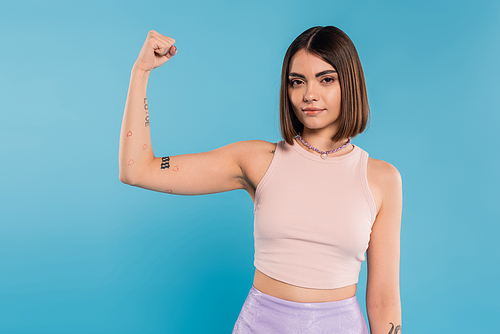 female power, brunette young woman with short hair, tattoos and nose piercing showing muscle on blue background, generation z, displeased, casual attire, strength