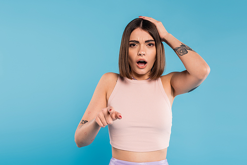 shocked face, brunette young woman with short hair, tattoos and nose piercing pointing at camera on blue background, generation z, displeased, casual attire, summer outfit