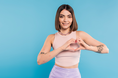 positivity and love, happy young woman with short hair, tattoos and nose piercing showing heart gesture with hands on blue background, generation z, cheerful, casual attire