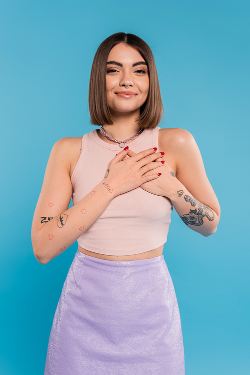 positivity and peace, happy young woman with short hair, tattoos and nose piercing holding hands near chest on blue background, generation z, cheerful, casual attire
