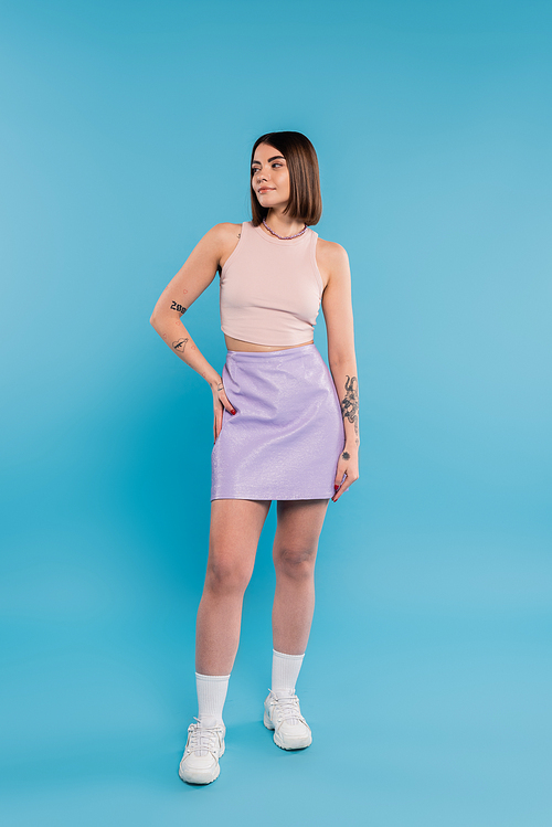 summer trends, brunette young woman with short hair in tank top, skirt and white sneakers posing on blue background, casual attire, gen z fashion, personal style, nose piercing, hand on hip