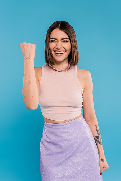 summer trends, positive young woman with short hair in tank top and skirt showing yes gesture on blue background, casual attire, gen z fashion, personal style, nose piercing, excitement