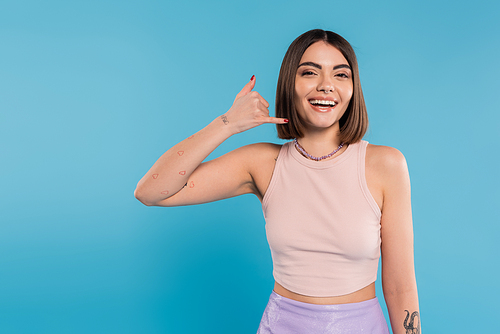 showing call me, happy young woman with short hair gesturing and looking at camera on blue background, casual attire, gen z fashion, personal style, nose piercing, positivity