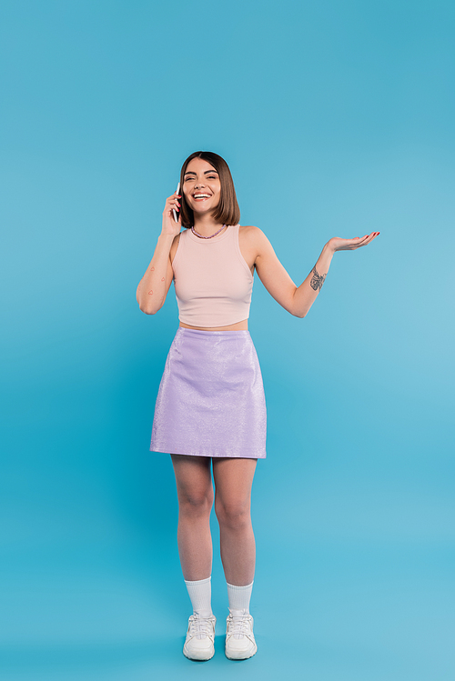 full length, phone call, cheerful young woman with short hair, tattoos and nose piercing gesturing while talking on smartphone on blue background, casual attire, gen z fashion, personal style
