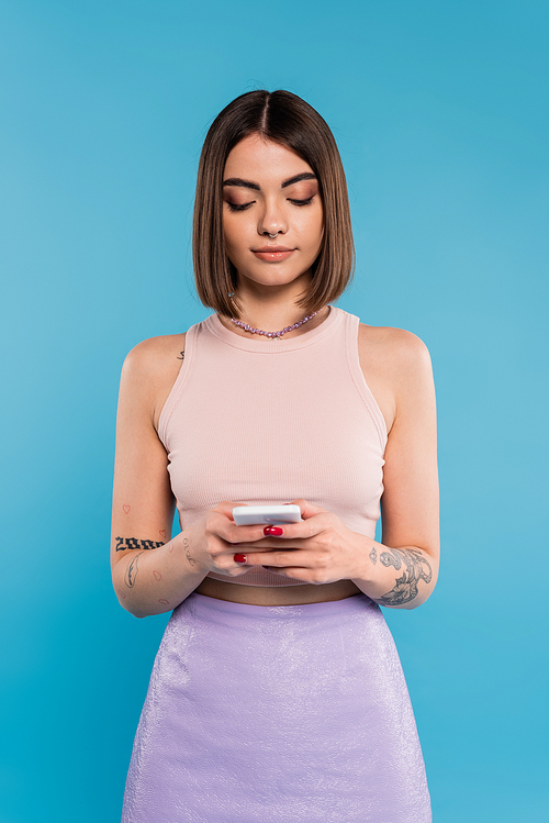 woman texting on smartphone, short hair, tattoos and nose piercing using mobile phone on blue background, casual attire, gen z fashion, personal style, everyday makeup