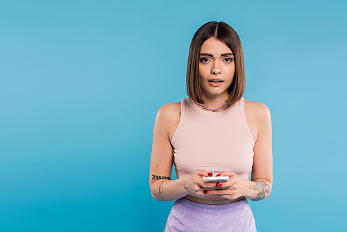 messaging on smartphone, shocked, young brunette woman short hair, tattoos and nose piercing using mobile phone on blue background, casual attire, gen z fashion, social media influencers