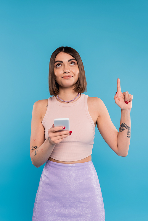 holding smartphone, young brunette woman short hair, tattoos and nose piercing pointing up on blue background, casual attire, gen z fashion, social media influencers