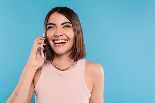 phone call, positivity, cheerful young woman with short hair, tattoos and nose piercing talking on smartphone on blue background, casual attire, gen z fashion, personal style