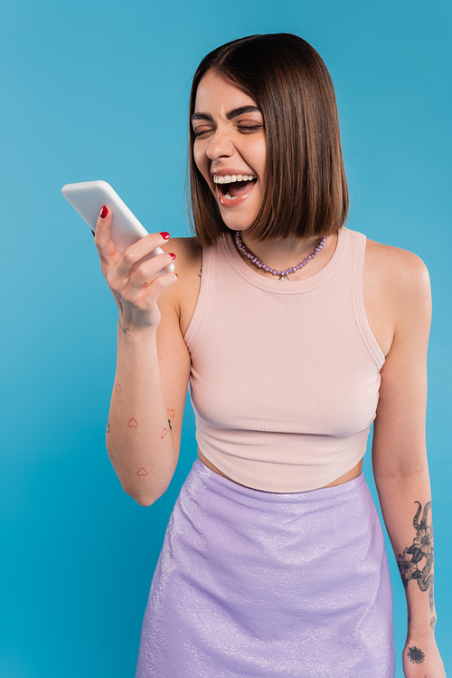 sending a message, excited young woman short hair, tattoos and nose piercing using mobile phone on blue background, casual attire, gen z fashion, social media influencers