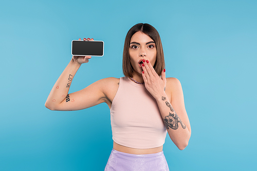 smartphone with blank screen, shocked young woman with short hair, tattoos and nose piercing holding mobile phone on blue background, gen z fashion, social media influencers, cover mouth