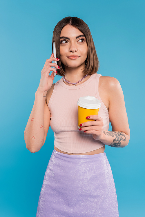 to go coffee, happy and tattooed young woman with short hair and nose piercing holding paper cup and talking on smartphone on blue background, generation z, summer trends, attractive, coffee culture