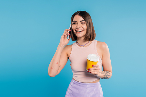 to go coffee, joyful and tattooed young woman with short hair and nose piercing holding paper cup and talking on smartphone on blue background, generation z, attractive, coffee culture