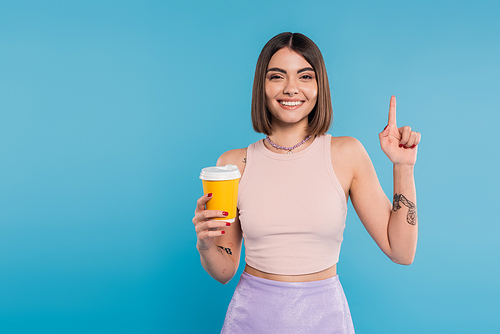 to go coffee, happy young woman with short hair, tattoos and nose piercing holding paper cup on blue background, generation z, summer trends, attractive, coffee culture, everyday style, pointing up