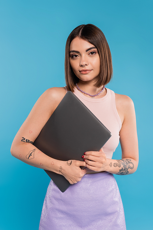 freelancer, brunette young woman with short hair, tattoos and nose piercing holding laptop on blue background, generation z, summer trends, attractive, remote work, everyday style