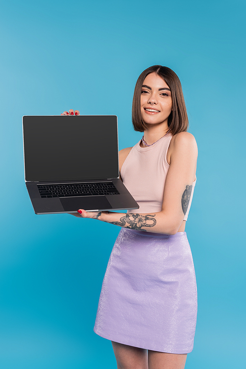 freelancer, cheerful young woman with short hair, tattoos and nose piercing holding laptop on blue background, generation z, summer trends, attractive, remote work, everyday style, blank screen