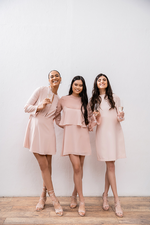 three multicultural bridesmaids, pretty women in pastel pink dresses holding glasses of champagne on grey background, cultural diversity, fashion, celebration, cheers