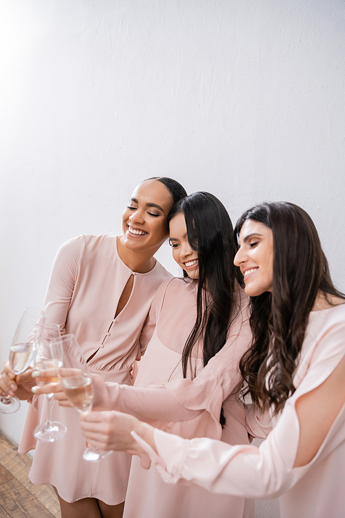 three multicultural bridesmaids, pretty women in pastel pink dresses clinking glasses of champagne on grey background, diversity, fashion, celebration, cheers, diverse races