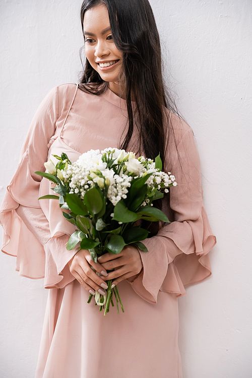 happy bridesmaid in pastel pink dress holding bouquet, asian woman with brunette hair on grey background, white flowers, special occasion, wedding, fashion, smile and joy