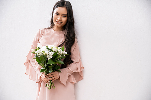 cheerful bridesmaid in pastel pink dress holding bouquet, asian woman with brunette hair on grey background, white flowers, special occasion, wedding, fashion, smile and joy, looking at camera