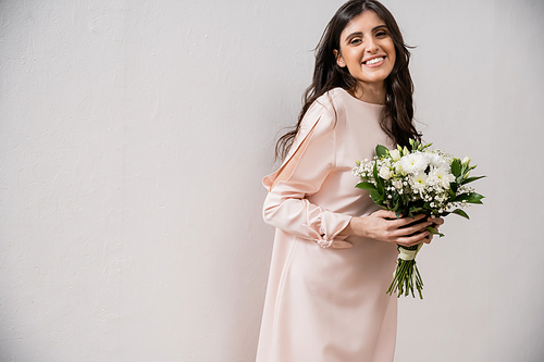 cheerful bridesmaid in pastel pink dress holding bridal bouquet, brunette woman on grey background, white flowers, special occasion, wedding, fashion, smile and joy, looking at camera