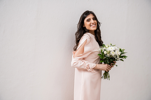 positive bridesmaid in pastel pink dress holding bridal bouquet, brunette woman on grey background, white flowers, special occasion, wedding, fashion, smile and joy, looking at camera