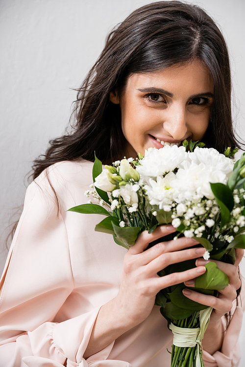 special occasion, happy bridesmaid in pastel pink dress holding bouquet, brunette woman on grey background, white flowers, wedding, fashion, smile and joy, looking at camera